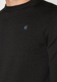 Schwarzer Strickpullover mit Rundhalsausschnitt und gerippten Bündchen. Verfügt über ein kleines blaues Logo auf der Brust, das einen strukturierten Stoff zeigt.