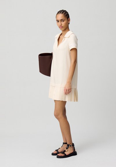 Vestido beige claro con cuello tipo camisa, mangas cortas y dobladillo plisado. La modelo lleva un bolso redondo de color marrón oscuro y sandalias negras.