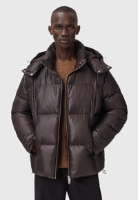 Mørkebrun puffjakke med hætte, båret over en brun strikket sweater, lynet halvvejs op, med hænderne i lommen, på en stående mandlig model.