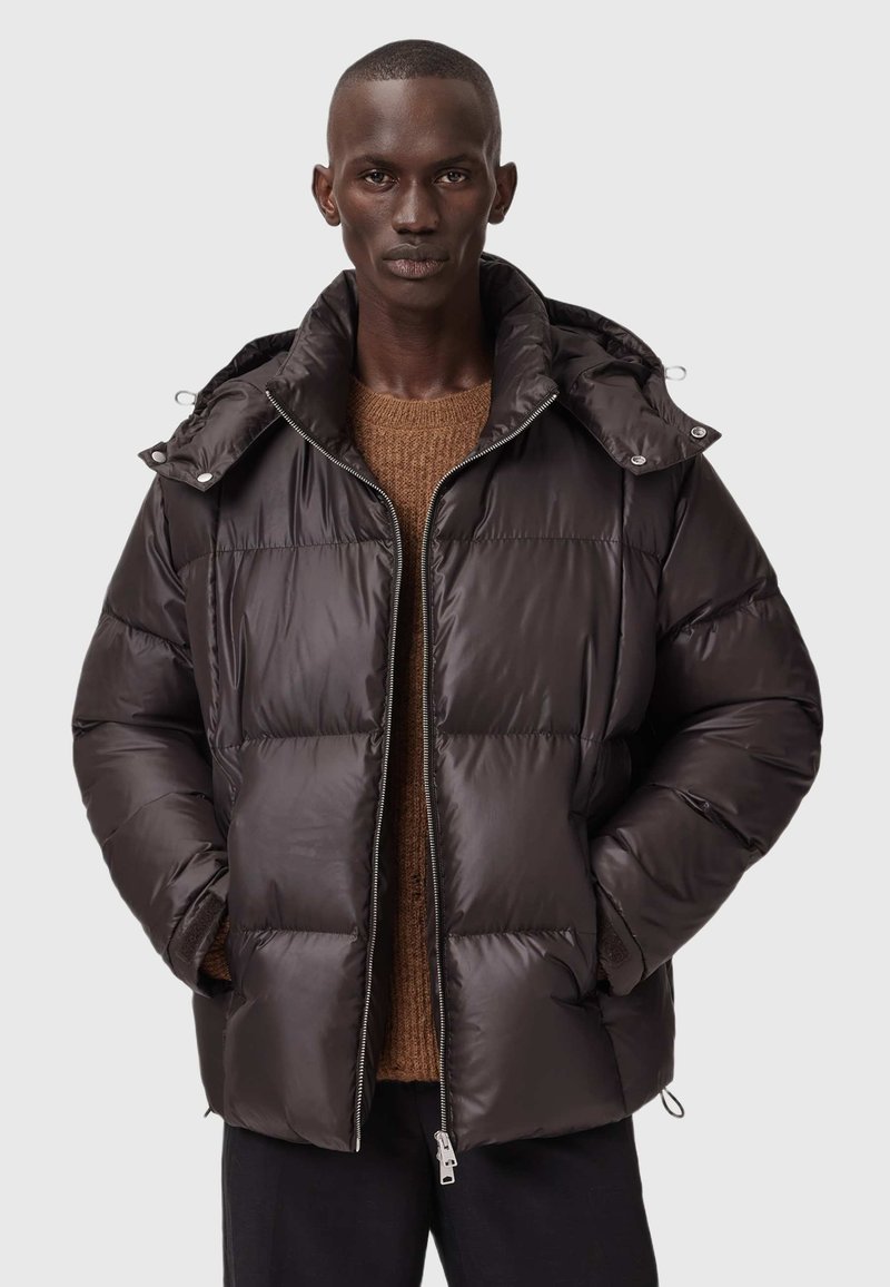 Mørkebrun puffjakke med hætte, båret over en brun strikket sweater, lynet halvvejs op, med hænderne i lommen, på en stående mandlig model.