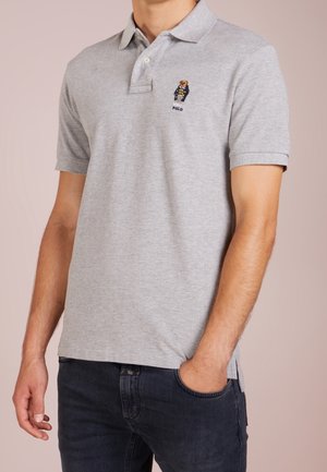 Mand iført en lysegrå polo med en lille broderet bjørnelogo og mørke jeans, med den ene hånd i lommen og neutral baggrund.