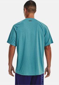 T-shirt sportowy z krótkim rękawem w kolorze teal, z teksturowanej tkaniny, okrągłym dekoltem i logiem na plecach. Noszony z fioletowymi szortami, z bocznym brandingiem.