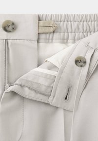 Gros plan sur la ceinture d’un pantalon beige montrant un bouton, une boutonnière, une fermeture éclair, un élastique réglable et les détails des coutures intérieures.