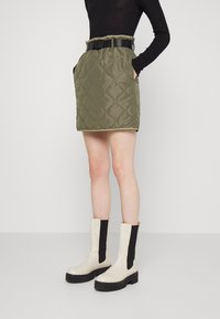 Jupe olive verte matelassée avec une ceinture noire, présentant des poches latérales et une coupe droite. Portée avec des bottines noires et crème.