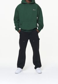 Felpa verde con cappuccio e tasca frontale, abbinata a pantaloni cargo neri con tasche laterali e dettagli in argento. Sneakers bianche completano il look.