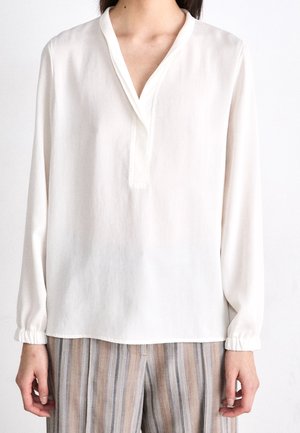 Frau trägt eine locker sitzende weiße Bluse mit langen Ärmeln, V-Ausschnitt und elastischen Bündchen, kombiniert mit beige-grau gestreiften Hosen.