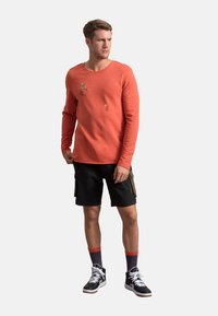 Camicia a maniche lunghe color corallo con collo a giro, caratterizzata da grafiche colorate, abbinata a pantaloni cargo neri e sneakers alte nere.