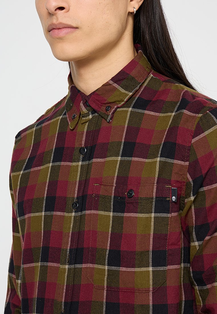 Chemise écossaise en rouge, vert et noir, fabriquée en tissu tissé. Elle présente un col boutonné, une poche avant et des boutons noirs.