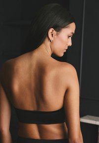 Czarny top bandeau noszony przez osobę, cechujący się smukłym designem i gładką teksturą. Odcień skóry jest ciepły, a tło ciemne i minimalistyczne.