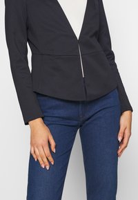 Marineblauw blazer met lange mouwen, gestructureerde pasvorm, diepe V-hals en een scherpe voornaad. Gecombineerd met donkerblauwe jeans, met een soepele denimtextuur.