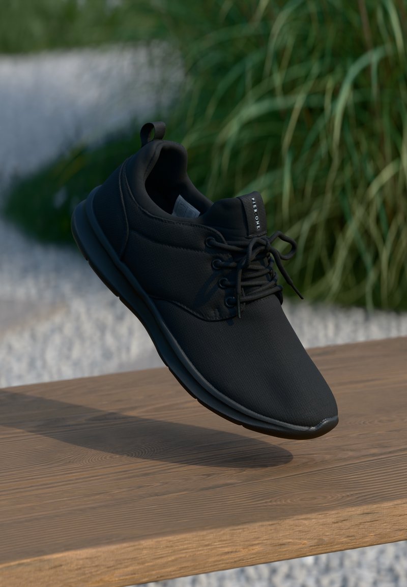 Baskets noires décontractées avec lacets, suspendues au-dessus d'une surface en bois avec des plantes vertes floues en arrière-plan.
