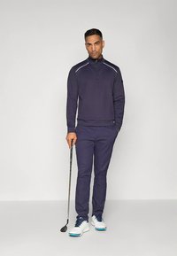 Tengerészkék cipzáros golfpulóver és hozzáillő nadrág. Sima anyag, fehér kontrasztvarrás. Modell golfütőt tart, fehér cipőt visel kék fűzőkkel.