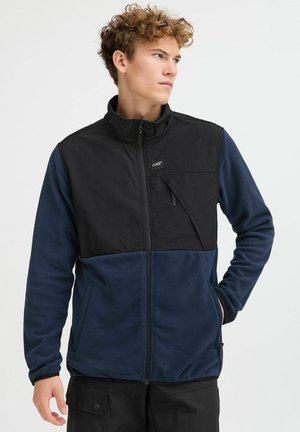 Veste en polaire noire et marine avec un col haut, des poches latérales zippées et une fermeture éclair à l'avant. Présente des textures de tissus contrastées.