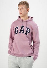 Pullover hoodie in soft lavender fabric with a front kangaroo pocket. Features large navy "GAP" lettering on the chest. Paired with grey sweatpants.
