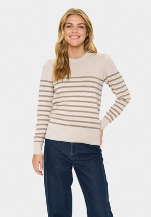 Smilende kvinde med langt blond hår iført en beige og brun stribet sweater og mørkeblå jeans, stående med den ene hånd på hoften.