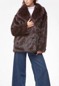 Cappotto in pelliccia sintetica marrone con un ampio colletto, maniche lunghe e una vestibilità morbida. Superficie testurizzata e morbida, abbinato a jeans blu a gamba larga.