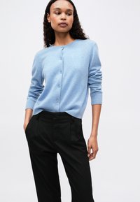 O femeie cu păr creț, îmbrăcată cu un cardigan butonat de un bleu deschis și pantaloni negri cu talie înaltă, stând cu mâinile în buzunare pe un fundal deschis.