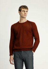 Brązowy sweter w zygzakowy wzór z długimi rękawami i ściągaczami. Noszony na szare spodnie, z okrągłym dekoltem i teksturowanym splotem.