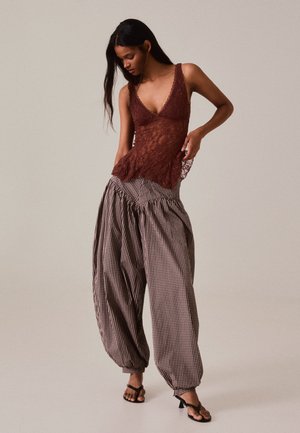 Femme portant un haut marron en dentelle sans manches, un pantalon ample à carreaux et des sandales noires à talons, debout devant un fond beige uni.