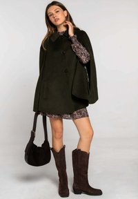 Cape en laine vert foncé avec un col, ornée de boutons noirs, associée à une robe à motif floral et des bottes hauteur genoux en suède marron.