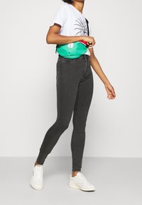 Bolso de cintura verde con cremallera y detalles rojos, llevado sobre una camiseta gráfica blanca, combinado con jeans ajustados grises y zapatillas blancas.