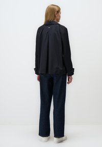 Czarna wodoodporna kurtka z plisą na plecach, z guzikami przy mankietach i subtelnym logo. Zestawiona z luźnymi niebieskimi dżinsami i białymi trampkami.