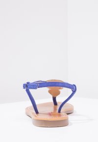 Blå mockasammet sandal med justerbar vristrem, platt brun sula och minimalistisk design. Har ett dekorativt element på toppen.