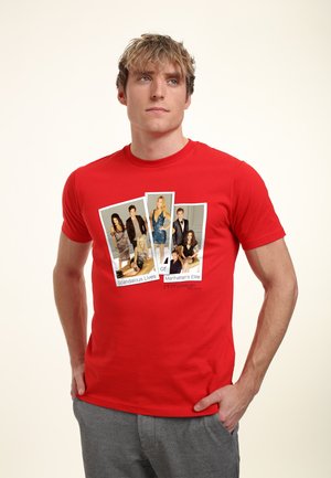 Young man wearing a red T-shirt with a print of six stylish people in formal and casual outfits arranged as three Polaroid photos.
