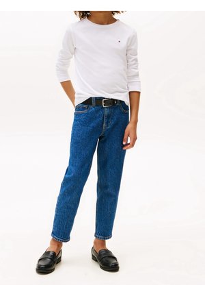 Person iført en hvid langærmet skjorte, blå denimjeans med sort bælte og sorte loafers står foran en ensfarvet baggrund.
