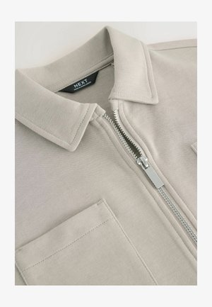 Jachetă cu fermoar de culoare gri deschis, având un guler plat și două buzunare frontale. Fabricată dintr-un material moale cu o textură netedă și fermoar metalic.