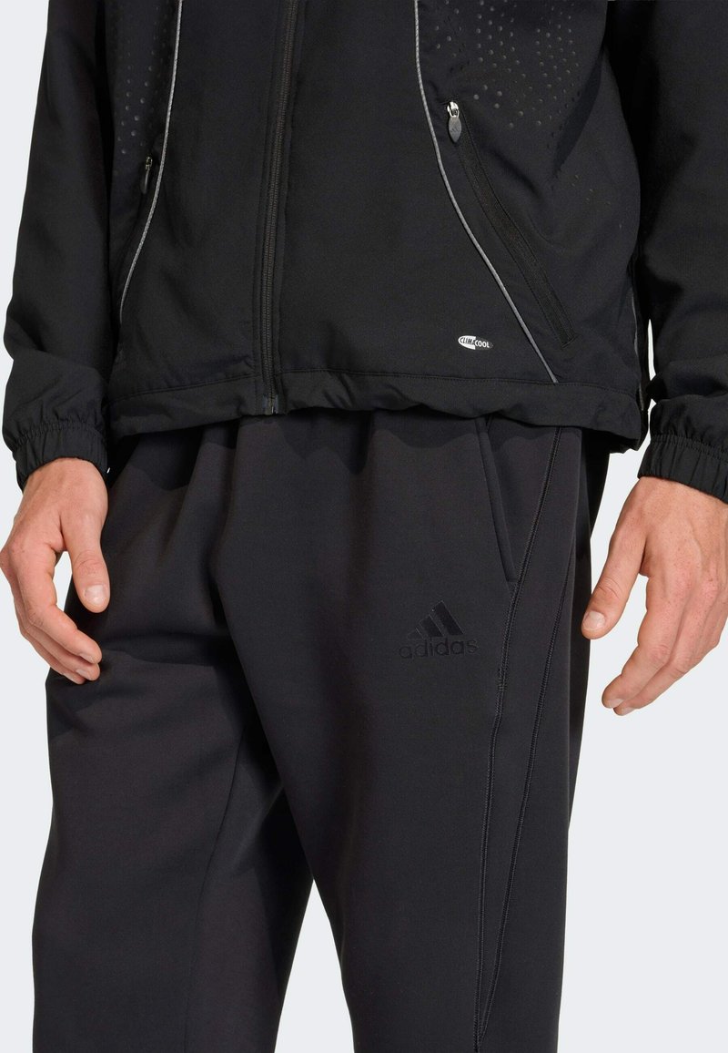 Uomo che indossa pantaloni da jogging neri Adidas con logo visibile e giacca nera con zip e etichetta ClimaCool, mani rilassate ai lati.