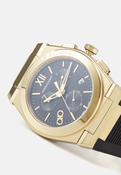 Gold-tone chronograph watch with a black textured dial, gold hands, Roman numerals, and a rubber strap. Date display at 3 o'clock position.