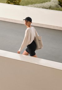 Világos bézs oversized pulóver hátul kivágással, fekete sort és szürke táska, sima felületen sétál egy minimalista környezetben.