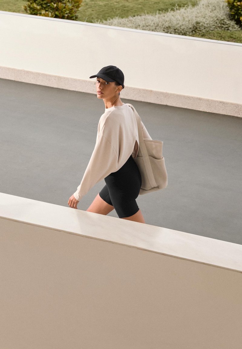 Világos bézs oversized pulóver hátul kivágással, fekete sort és szürke táska, sima felületen sétál egy minimalista környezetben.