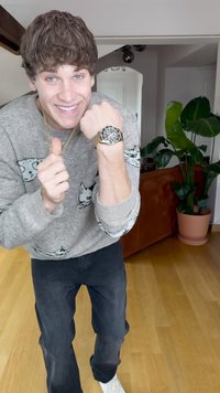 Sølv ur med mørk urskive og guld accenter, båret på en lys hudfarvet mands håndled, ved siden af en grå sweater med katte mønstre.