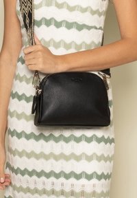 Borsa a tracolla in pelle nera con forma arrotondata, dotata di chiusura a zip e dettagli in metallo argentato. Indossata sulla spalla con una tracolla decorata.