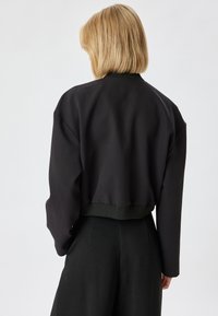 Lyhyt musta pom jacket, jossa on ribbaitut helmata ja hihansuut. Takapuolella näkyvät yksinkertaiset muotoiluelementit. Pinta on sileä ja malli väljä.