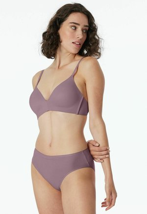 Woman with curly hair wearing mauve wireless bra and matching briefs, looking to her left, holding her right arm with left hand.
