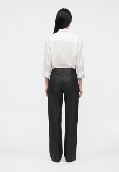 Personne aux longs cheveux noirs, portant une chemise blanche à manches longues et un pantalon en cuir noir, debout de dos sur un fond uni.
