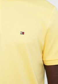 Chemise jaune à manches courtes avec un petit logo rectangulaire comportant des rayures rouges, blanches et bleu foncé sur la poitrine.