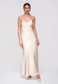 Femme portant une robe nuisette en satin crème avec des bretelles tressées et des escarpins crème pointus, debout devant un fond clair uni.