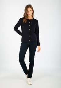 Granatowy kardigan z guzikami na przodzie, w połączeniu z ciemnymi spodniami i białymi trampkami. Gładka tekstura, okrągły dekolt i subtelny detal z logo.