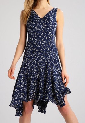 Hihaton tummansininen mekko, jossa valkoinen kukkakuvio, V-aukko ja epäsymmetrinen helma, päällä seisova henkilö yksiväristä taustaa vasten.