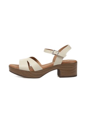 Sandalia de cuero beige con tacón de bloque de madera, correa ajustable en el tobillo con hebilla y dos tiras anchas delanteras que se cruzan sobre el pie.
