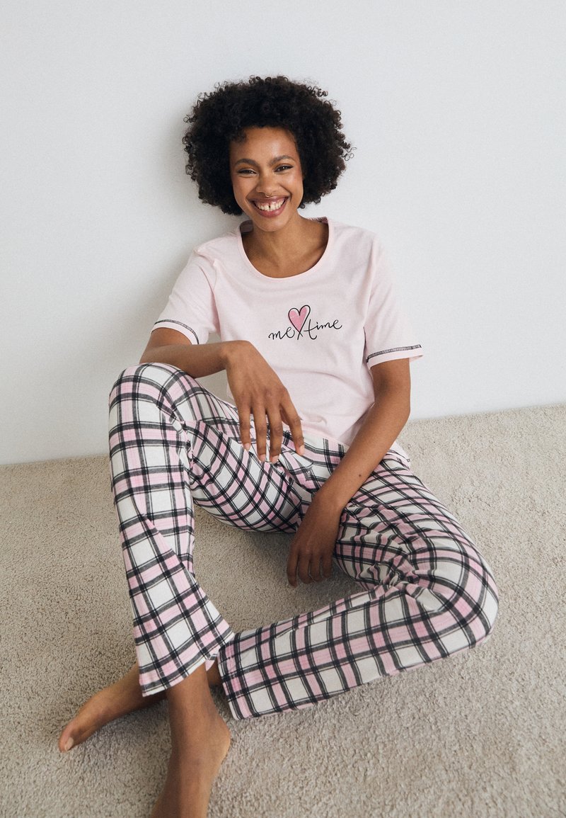 Camiseta de algodão rosa com o texto "me time", combinada com pijamas flare xadrez preto e rosa, exposta num fundo de carpete claro.