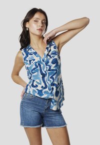 Camisa sin mangas azul y blanca con un patrón de ondas, que incluye un cuello, bolsillos frontales y cierre de botones, combinada con pantalones cortos de mezclilla.