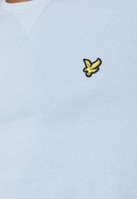 Světle modrý svetr vyrobený z pletené látky, s kulatým výstřihem a malým vyšitým logem žlutého ptáčka na levé hrudi.