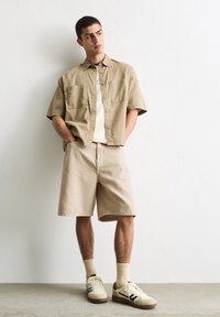 Beige Kurzarmhemd mit zwei Vordertaschen, kombiniert mit passenden beigen Shorts. Getragen mit cremefarbenen Sneakers und hellgelben Socken.