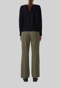 Schwarzer Strickpullover mit geripptem Saum, kombiniert mit olivgrünen, plissierten, weiten Hosen. Schwarze Schuhe runden das Outfit ab.