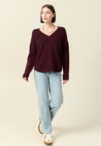 Bordeauxstickad tröja med V-ringning, avslappnad passform och långa ärmar, kombinerad med ljusblå jeans och vita sneakers med bruna detaljer.